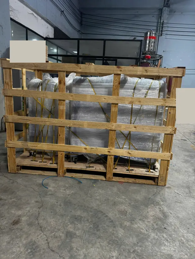 Wrapped machinery secured within a wooden frame; background labels and vehicle plates concealed